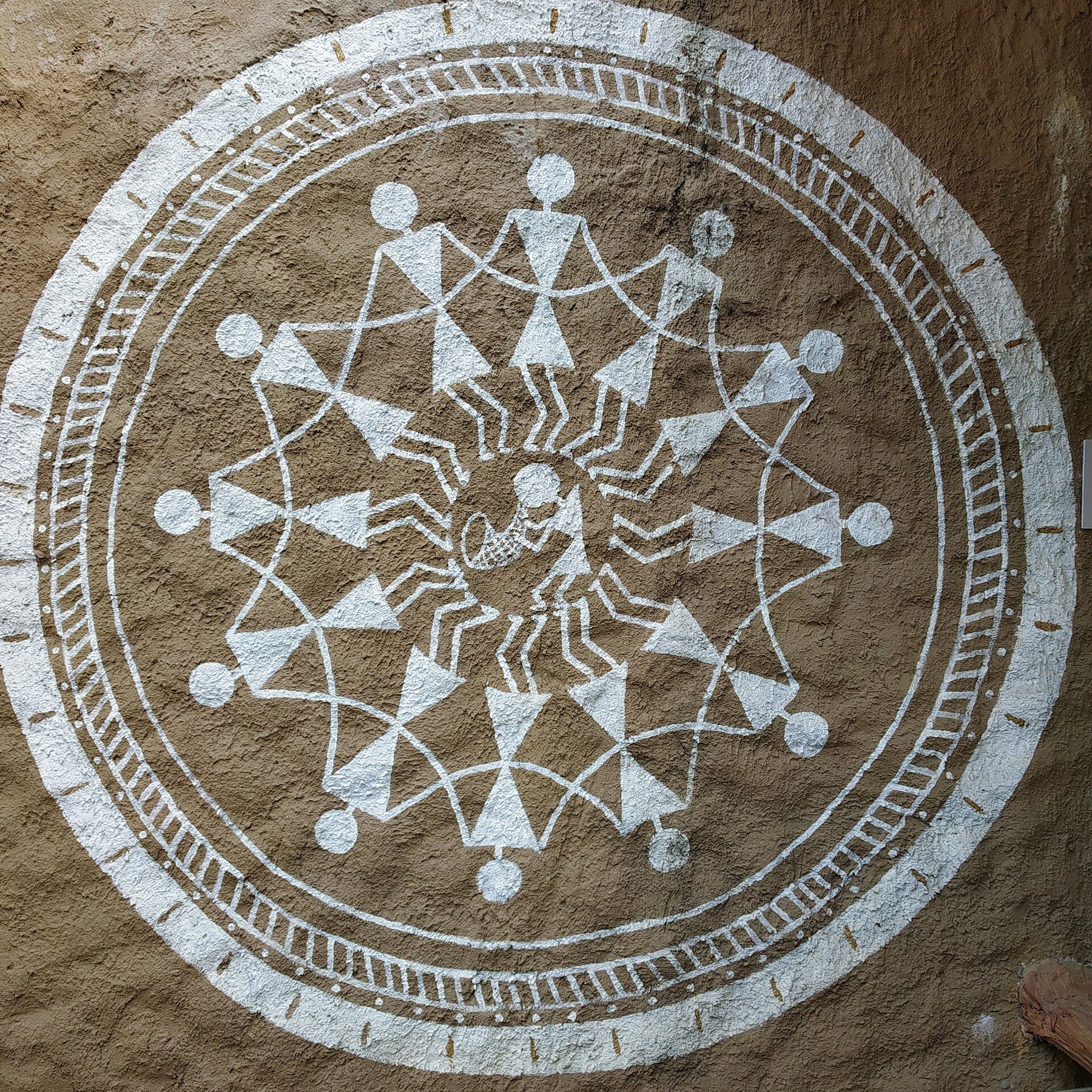 A Warli painting in white pigment on a brown ground, showing stylised figures, animals and a tree